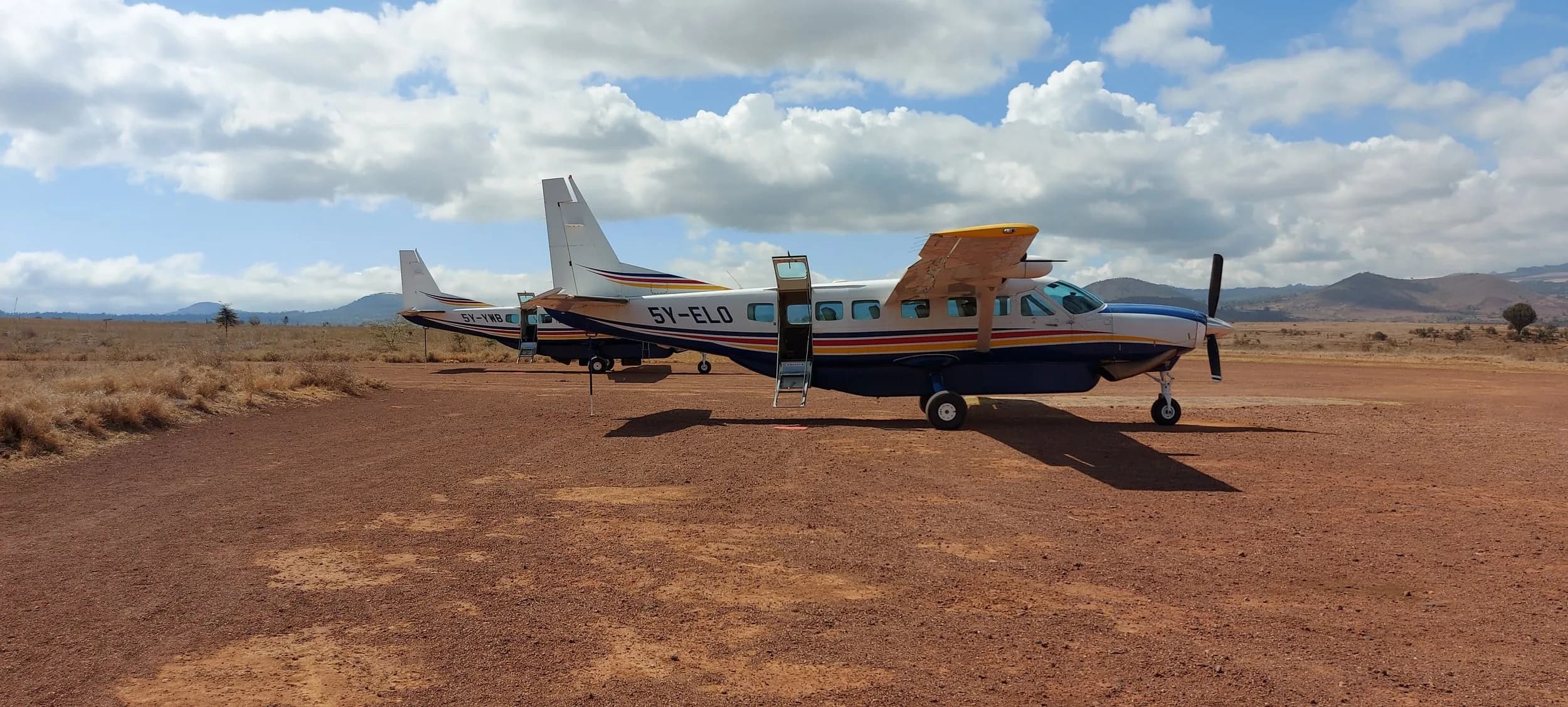 5Y-ELO and 5Y-YWB side by side on a red murram strip, 5Y-ELO's cargo door open